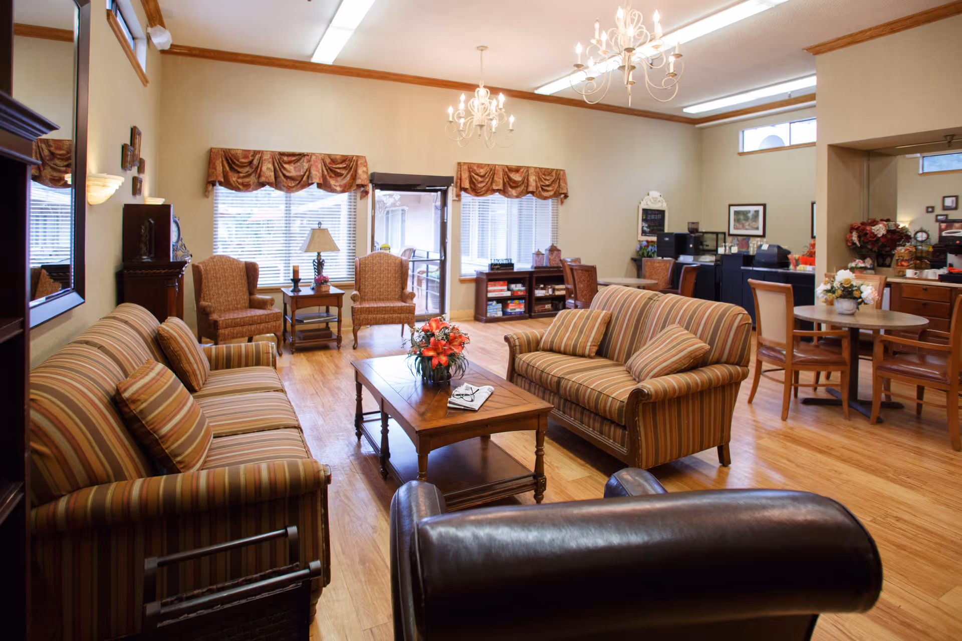 A cozy living room area in a senior living facility featuring two striped sofas, two armchairs, a wooden coffee table with a flower arrangement, and a leather chair in the foreground. The room has hardwood floors, large windows with valances, a chandelier, and a small dining table with chairs in the background. There are shelves with books and decorative items along the walls.