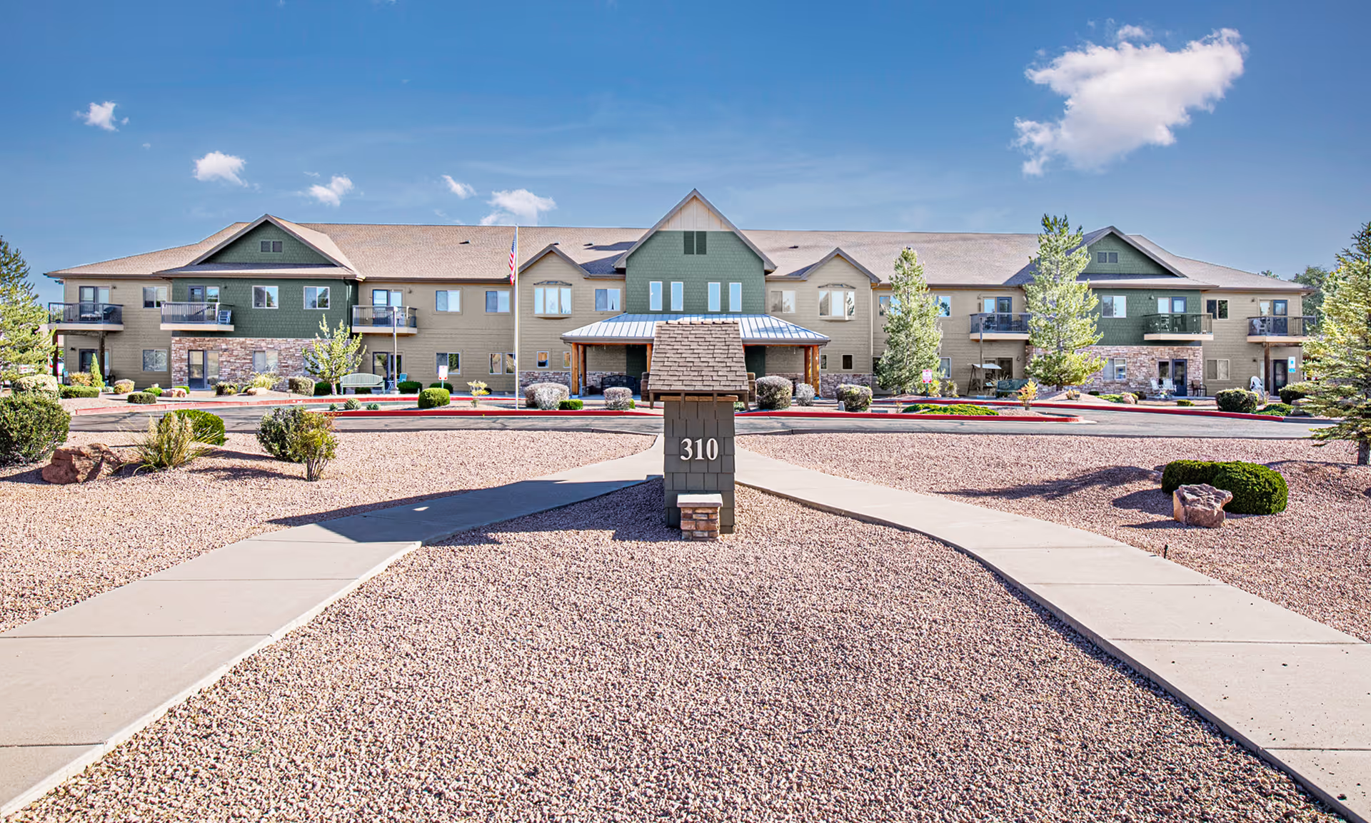 Front exterior view of a two-story retirement living facility with beige and green siding, balconies, and a central entrance. The landscaping includes gravel, small bushes, and trees under a clear blue sky.