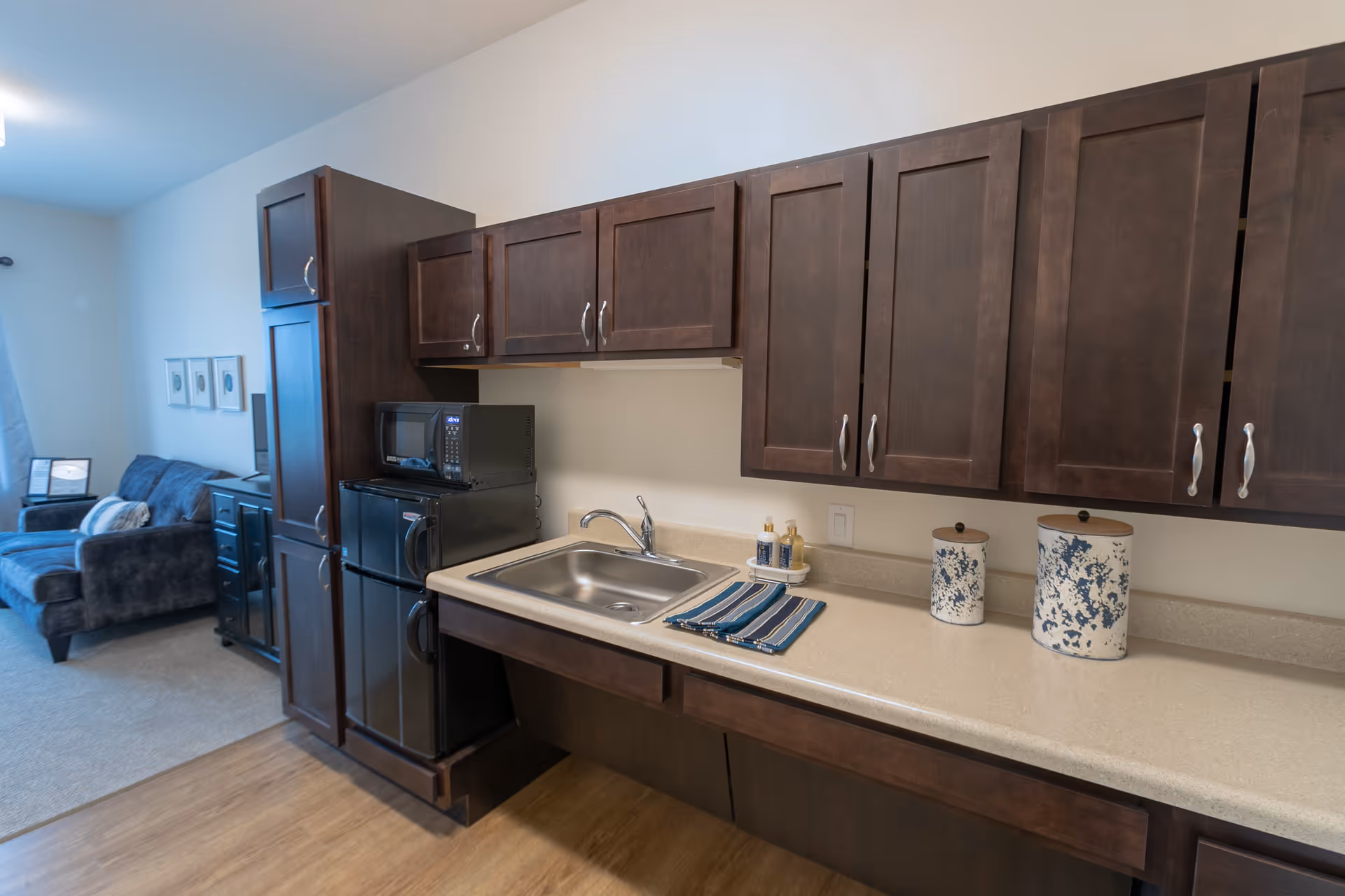 Interior view of a senior living facility kitchen area with dark wood cabinets, a small black refrigerator, a microwave, a stainless steel sink, and a countertop with two decorative canisters and a striped towel. In the background, a living room area with a dark upholstered sofa and framed pictures on the wall is visible.