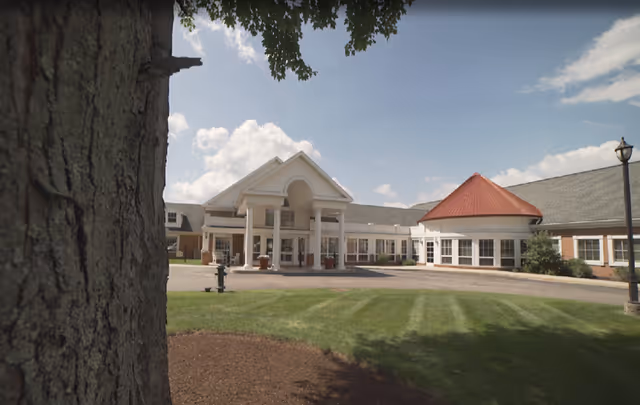 Exterior view of Grande Village Villas senior living facility showing a large building with a white entrance featuring columns and a peaked roof, a round section with a red conical roof, green lawn, a tree trunk in the foreground, and a partly cloudy sky.