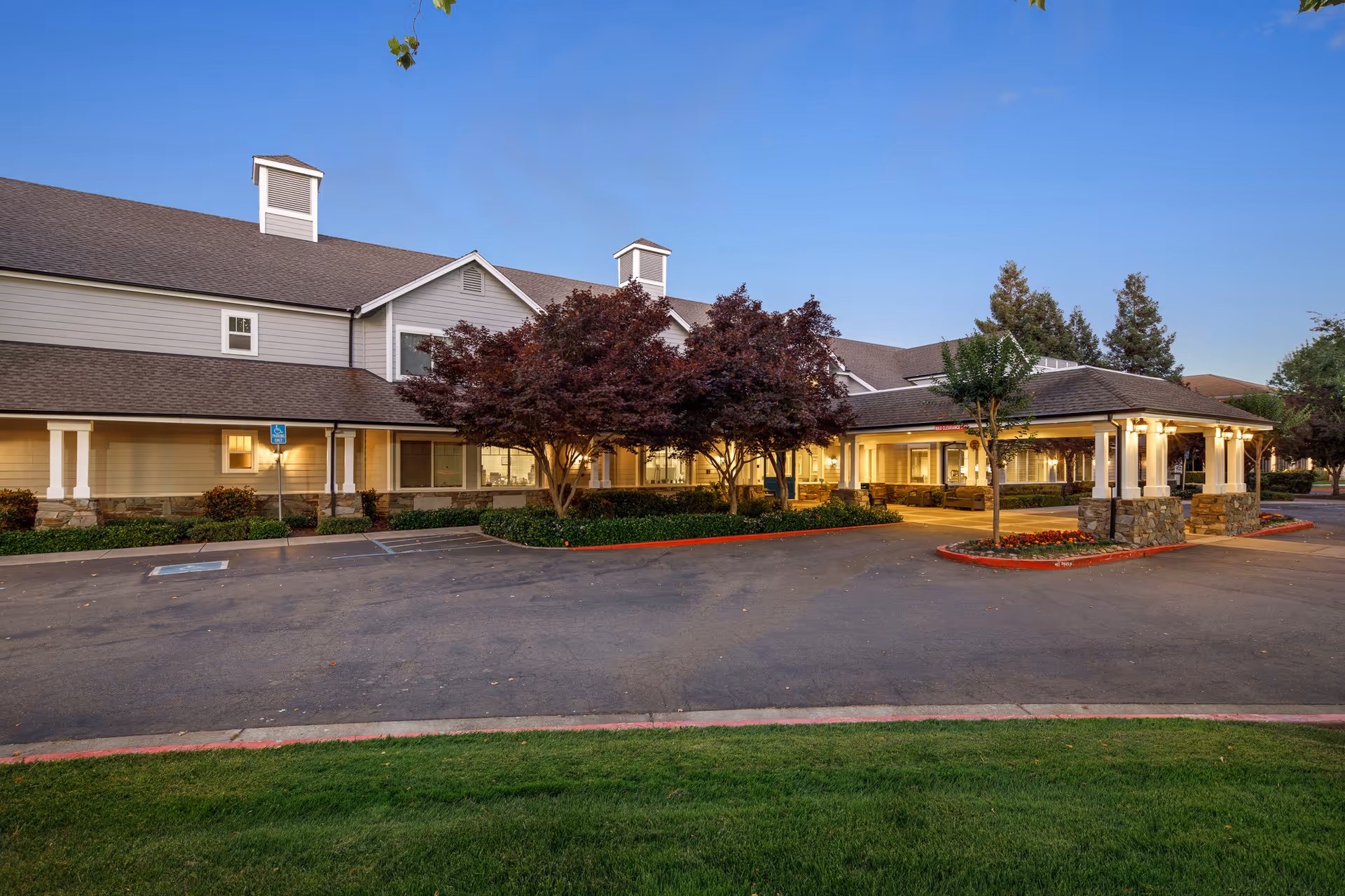 Exterior view of Oakmont Of Brookside senior living facility during dusk, showing a large building with beige siding, a covered entrance supported by stone pillars, landscaped greenery, and a parking area with a handicap parking sign.