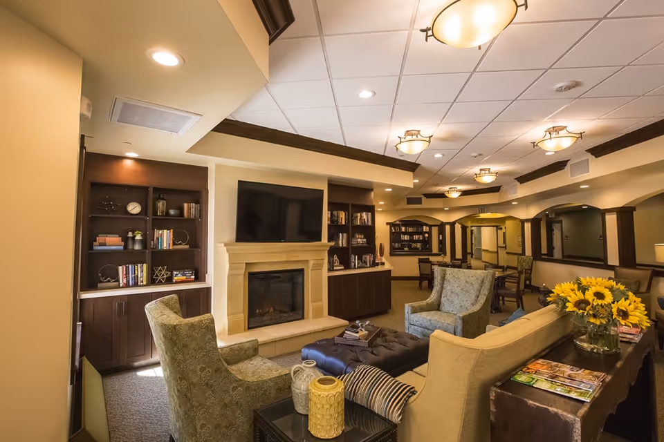 Cozy senior living common area with sofas and armchairs facing a fireplace and wall-mounted TV, with bookshelves and dining tables in the background.