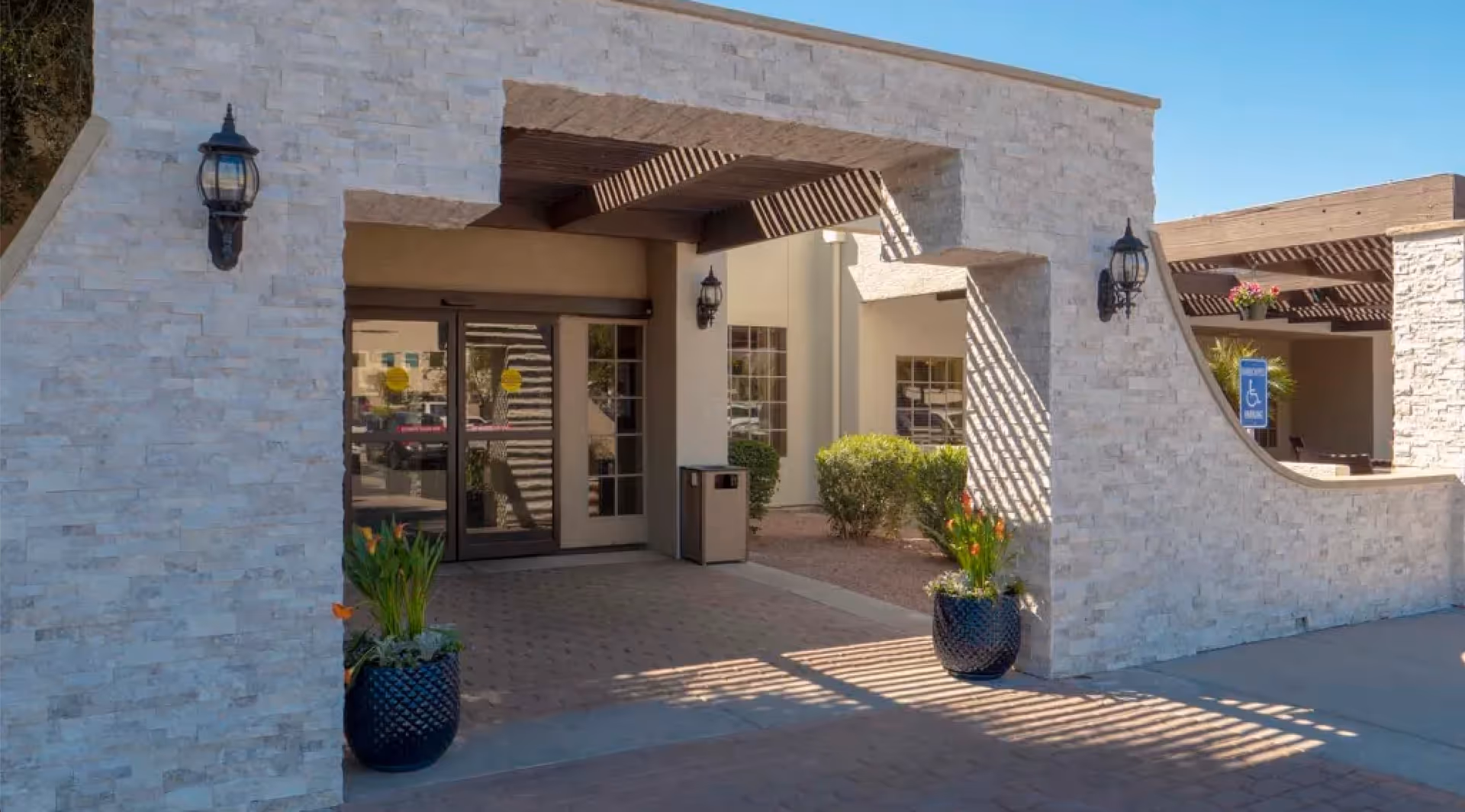 Entrance of a building with a stone facade and a covered walkway supported by wooden beams. There are two large planters with green plants and orange flowers on either side of the entrance. The entrance has glass double doors, and there are wall-mounted lantern-style lights on the stone walls. A handicap parking sign is visible to the right near some bushes.