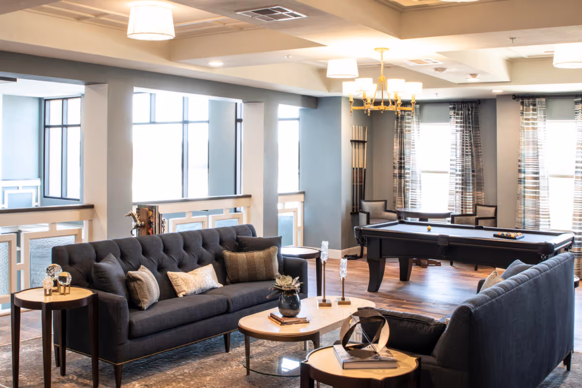 A bright and spacious common area with two dark gray sofas facing each other, a coffee table with decorative items, a side table, and a pool table in the background. Large windows with patterned curtains allow natural light to fill the room, and a chandelier hangs from the ceiling.