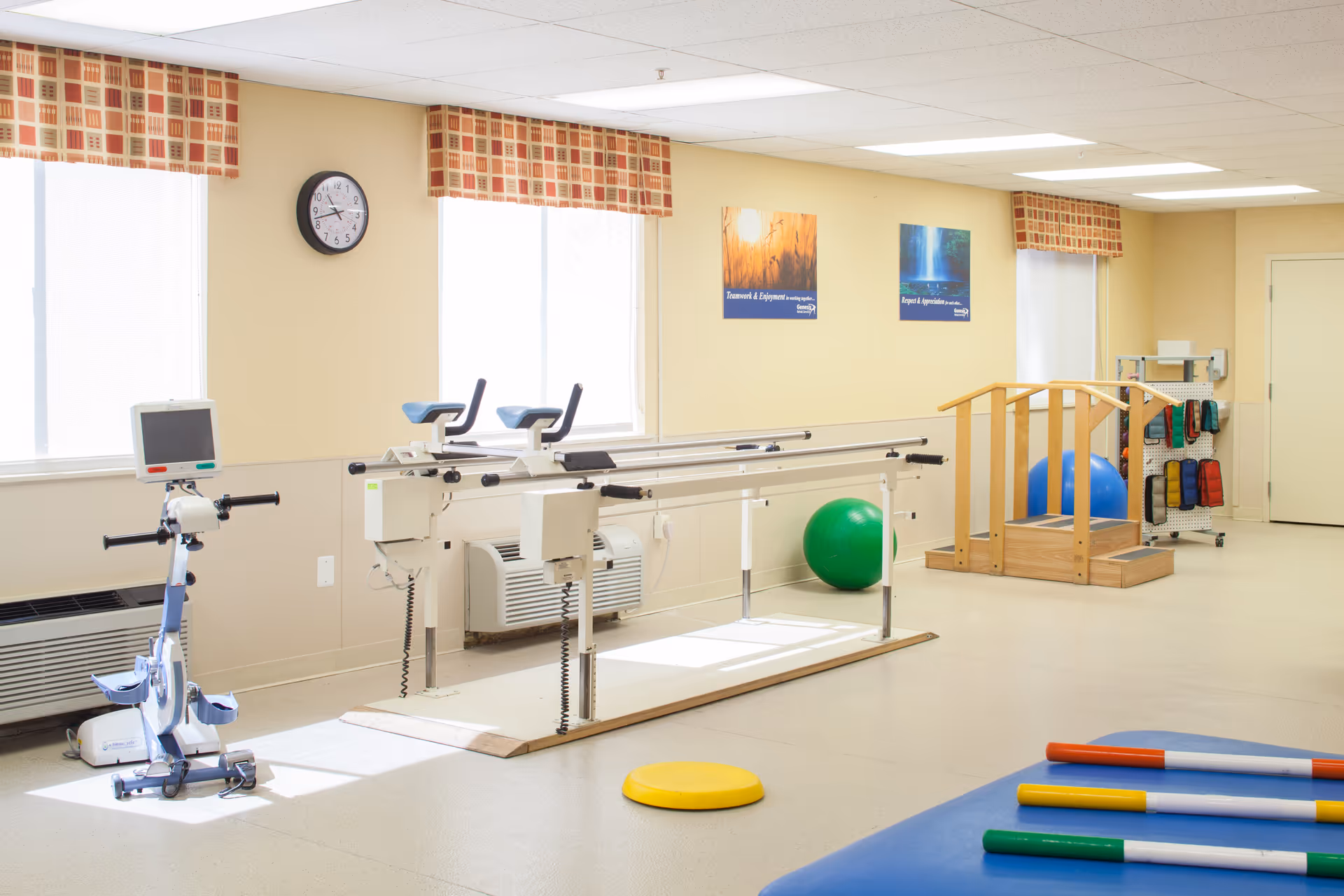 Bright rehabilitation room with parallel walking bars, an exercise bike, therapy balls, and a small stair simulator.