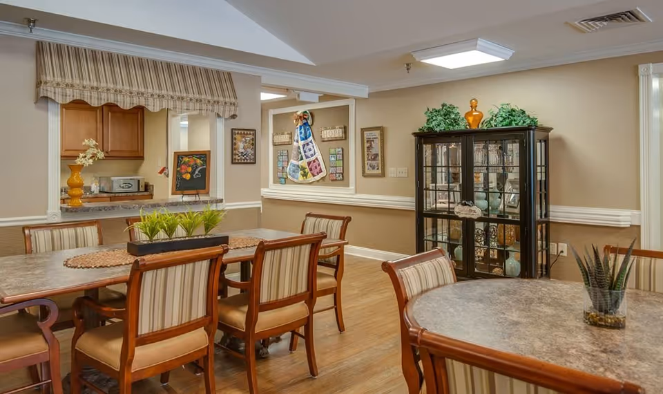 A dining room with several tables and striped-upholstered wooden chairs, a glass display cabinet, and a kitchen pass-through window.