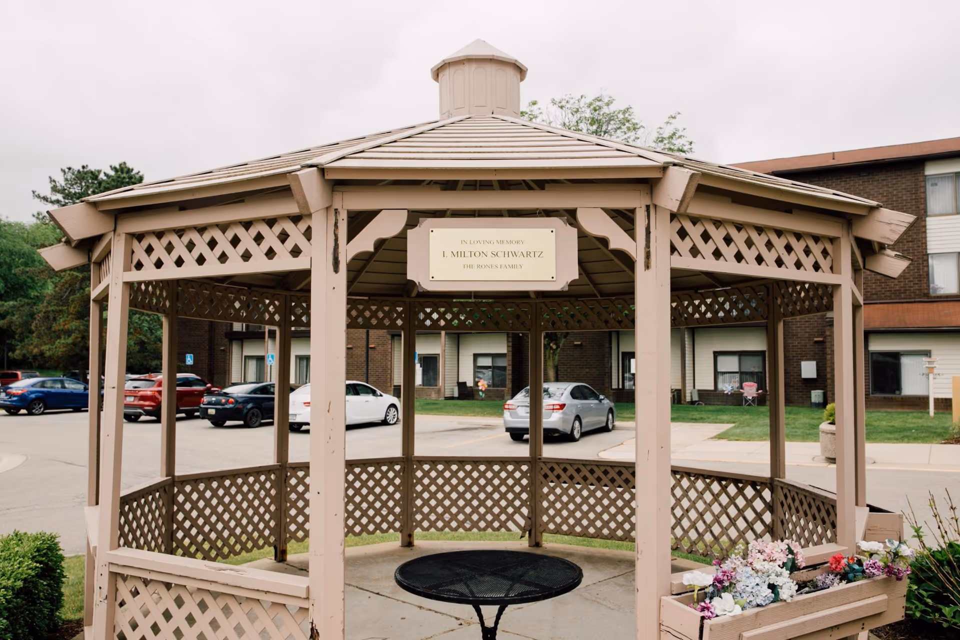 A beige wooden gazebo with lattice design located outdoors in front of a senior living facility building. There is a black round metal table inside the gazebo and a planter box with colorful flowers attached to the railing. Several parked cars and a brick building with windows are visible in the background. A sign inside the gazebo reads 'In Loving Memory L. Milton Schwartz The Bone's Family.'