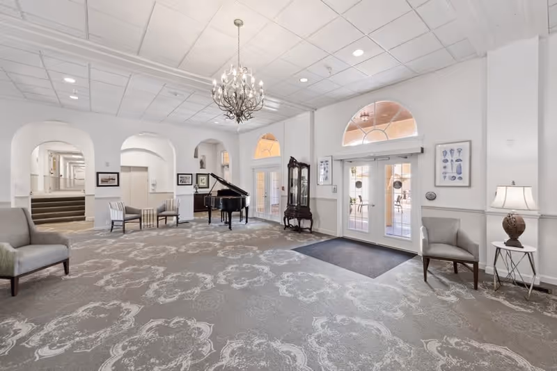 A spacious and elegant senior living facility common area with a grand piano, chandelier, patterned carpet, several chairs, a lamp on a side table, and large glass doors leading to an outdoor patio. The walls are white with arched doorways and framed artwork.