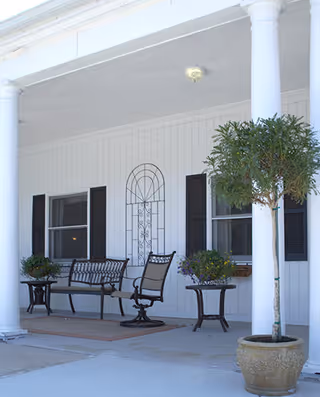 Covered front porch with white columns, bench seating, potted plants and windows with shutters.