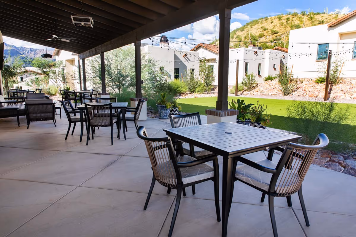 Outdoor covered patio area with multiple black metal tables and chairs arranged for seating. The patio overlooks a green lawn with desert landscaping and white buildings in the background under a blue sky with some clouds. String lights are hung across the patio ceiling.