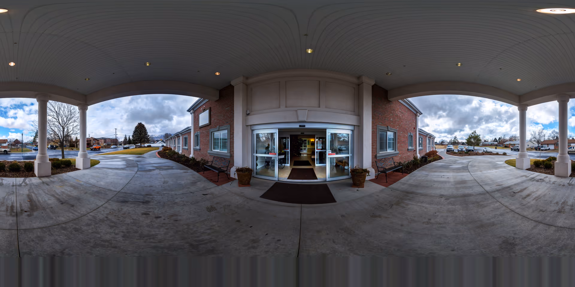Covered entrance area of a brick building with automatic sliding glass doors. There are benches and potted plants on either side of the entrance. The driveway curves around the entrance, and a parking lot with several cars is visible in the background under a partly cloudy sky.