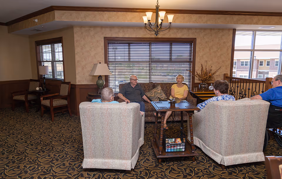 A group of elderly people sitting and chatting in a cozy senior living facility common area with patterned carpet, upholstered chairs, a sofa, a wooden coffee table, and large windows with blinds letting in natural light.