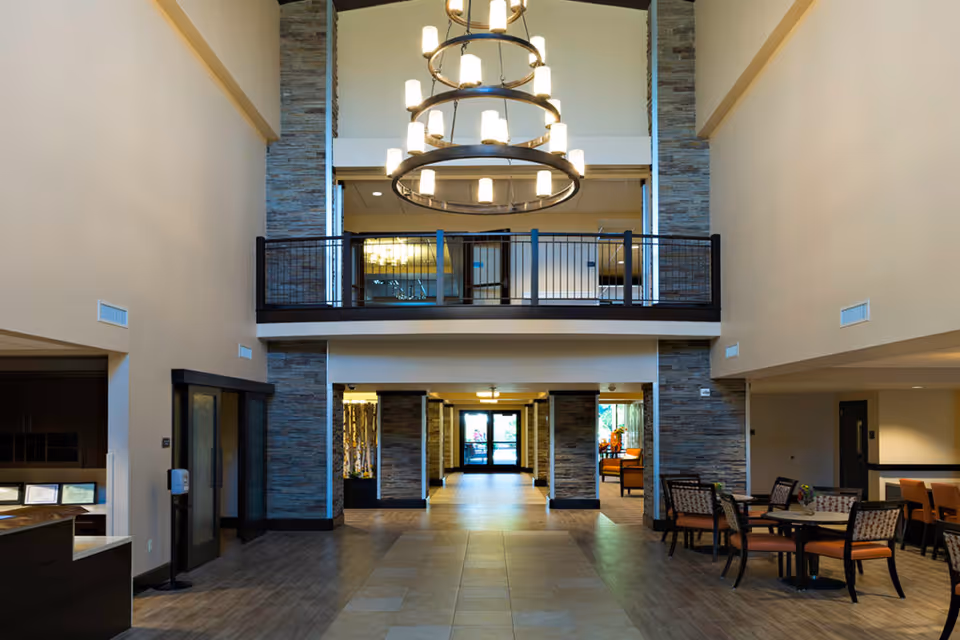 Spacious interior lobby area of The Ridge Cottonwood with high ceilings, large circular chandelier, stone pillars, and seating areas with tables and chairs. There is a balcony overlooking the lobby and a hallway leading to glass doors at the far end.