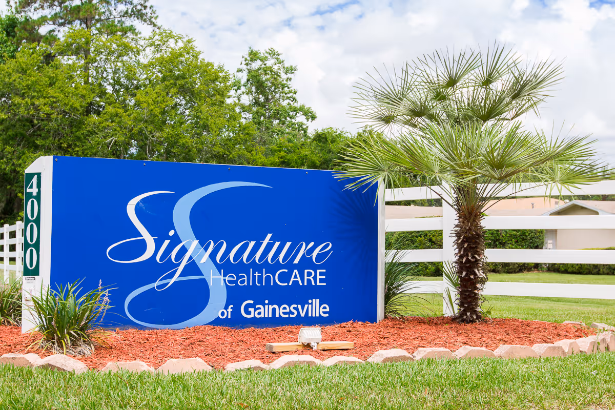 Outdoor view of a blue sign for Signature HealthCARE of Gainesville surrounded by landscaping with red mulch, green grass, a small palm tree, and other plants, with a white fence and trees in the background under a partly cloudy sky.