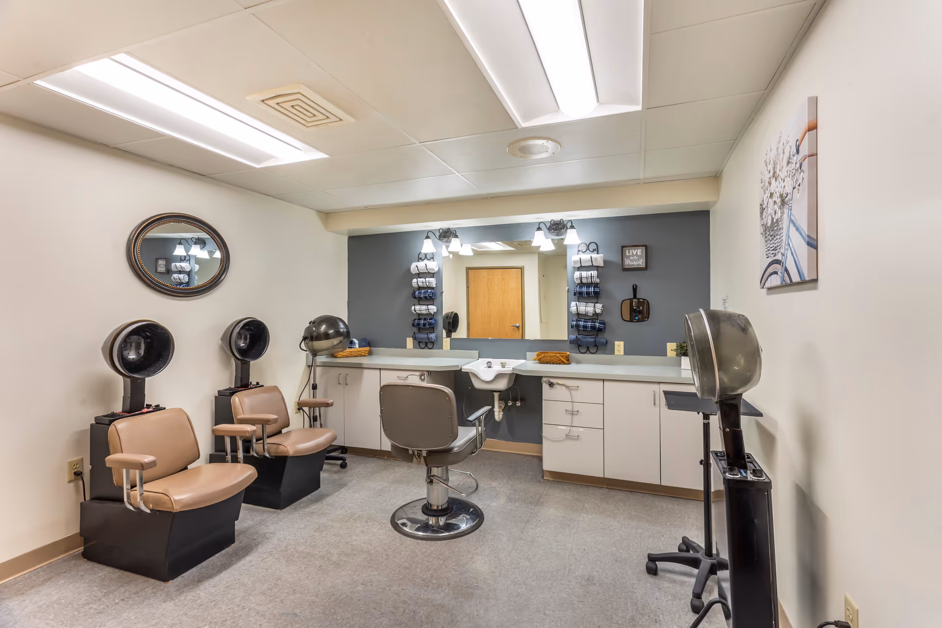 Interior view of a hair salon area in a senior living facility with three vintage hair drying chairs, a salon chair in front of a large mirror, a countertop with cabinets, and hair care products organized on the wall. The room has fluorescent ceiling lights and neutral-colored walls with a decorative picture and a small sign.