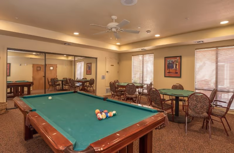 A recreational room with a green pool table in the foreground and several round tables with chairs in the background. The room has beige walls, a ceiling fan, large windows with blinds, and a framed picture on the wall. There is a large mirror on one side reflecting the room.