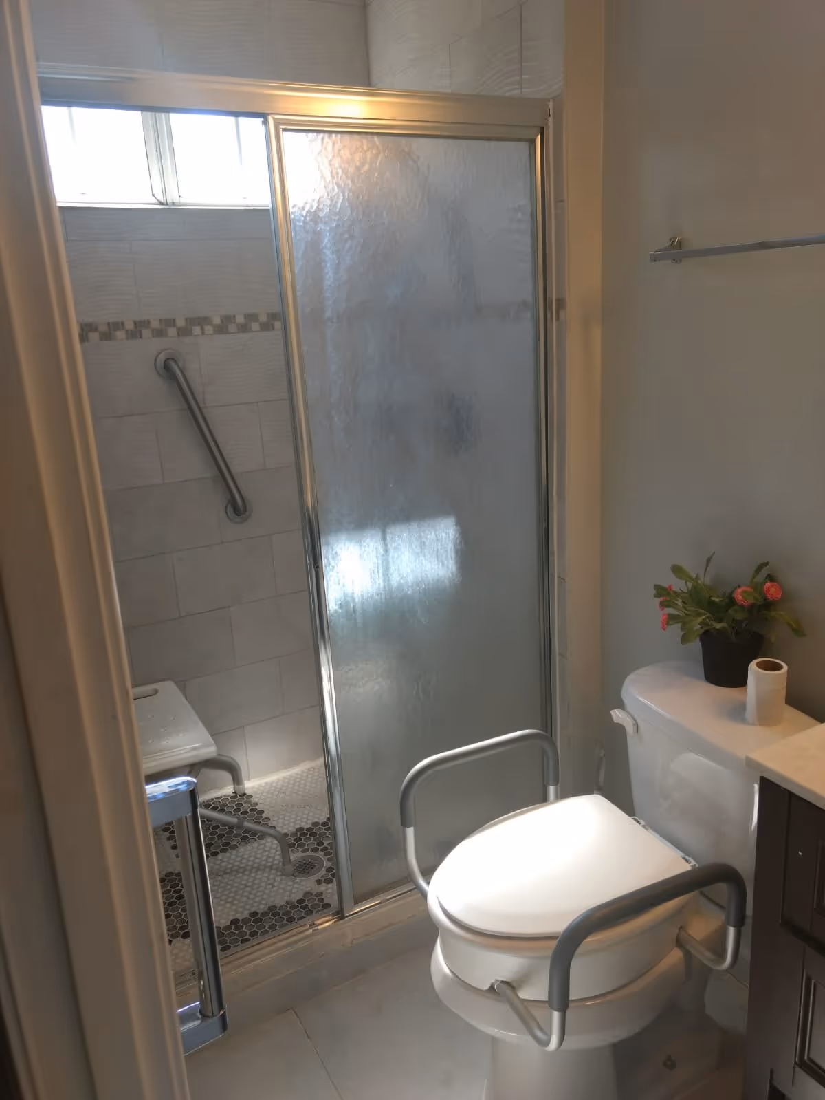 A bathroom with a toilet equipped with safety rails, a roll of toilet paper, and a small potted plant on the toilet tank. There is a shower area with a sliding frosted glass door, a grab bar on the tiled wall, and a shower chair inside. The bathroom has light-colored tiles and a towel rack on the wall.
