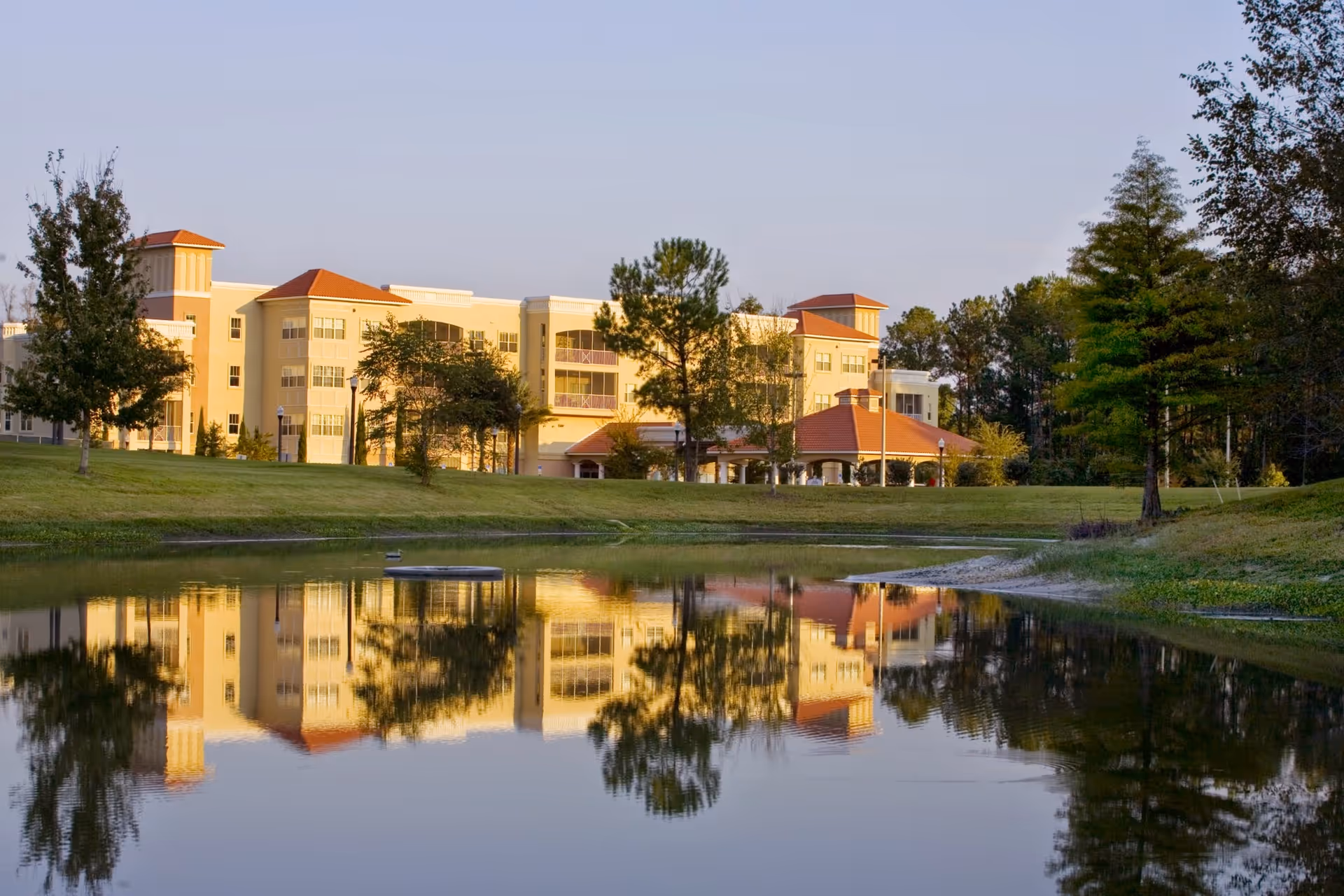A large senior living facility building with beige walls and red roofs, reflected in a calm pond in the foreground, surrounded by green grass and trees under a clear sky.