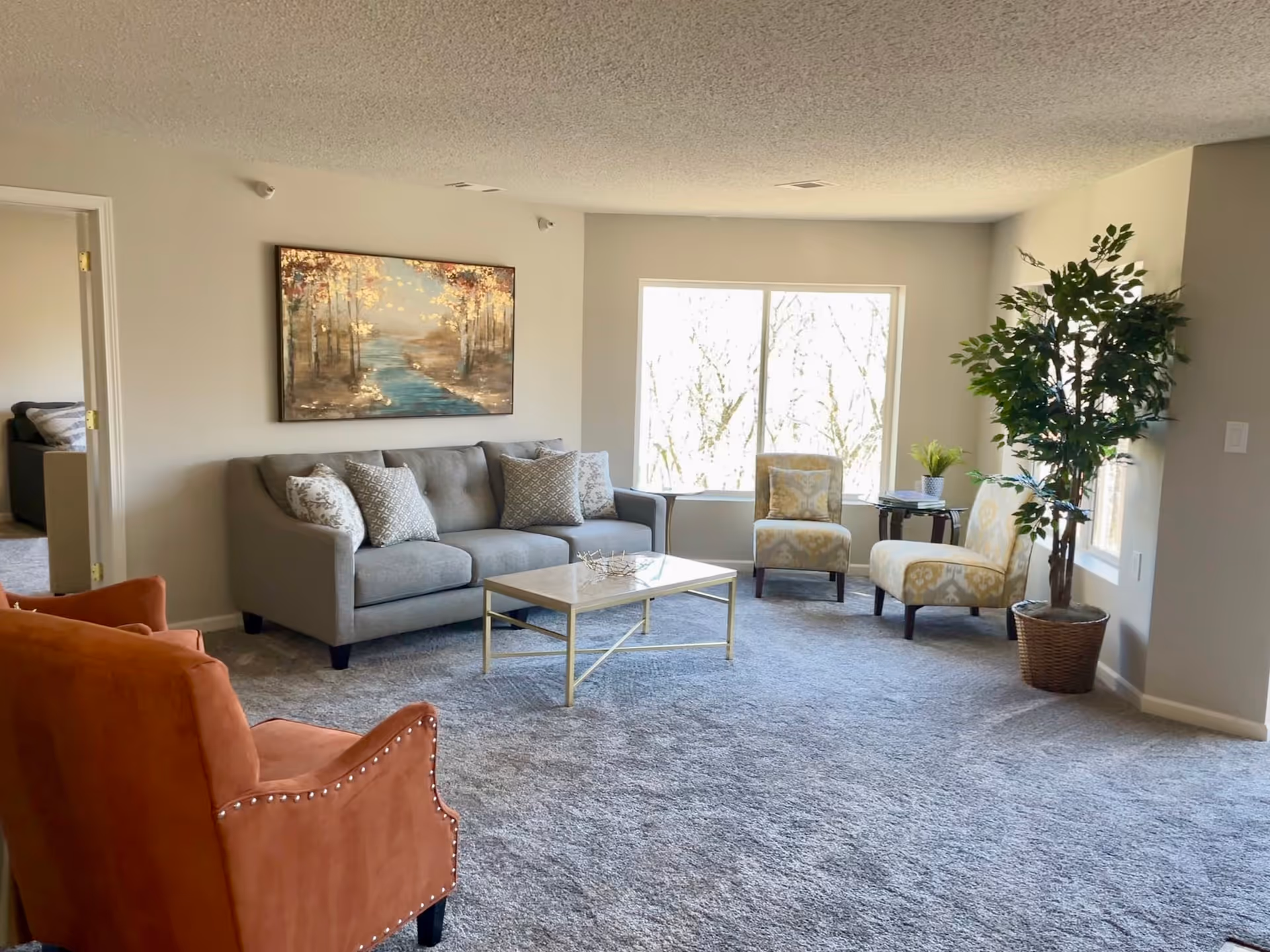 A bright living room with a gray sofa adorned with patterned pillows, two beige patterned chairs, and an orange armchair. A rectangular coffee table with a light-colored top sits in the center on a gray carpet. A large window lets in natural light, and a potted plant is placed near the window. A painting of a forest scene hangs on the wall above the sofa.
