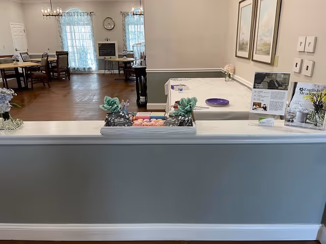 Interior view of a senior living facility dining area with wooden floors, several tables and chairs, large windows with sheer curtains, and decorative plants and flowers on a counter in the foreground. There are informational signs and framed artwork on the walls.