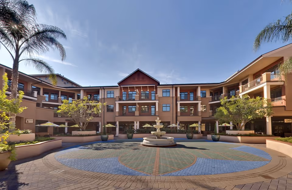 Outdoor courtyard area of Walnut Village Orange County Retirement Community featuring a central fountain, surrounded by potted plants, trees, and a three-story building with balconies and windows under a clear blue sky.