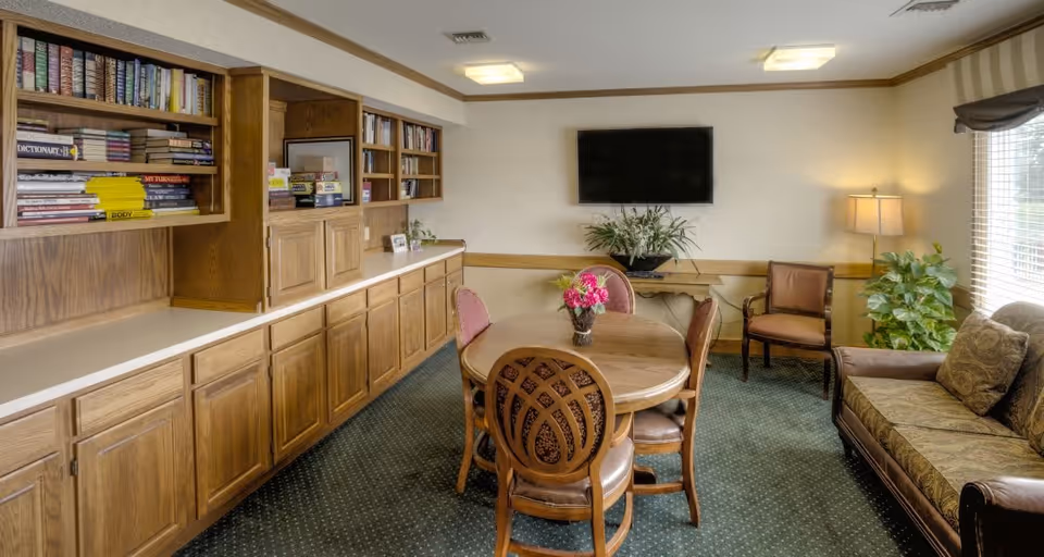 A cozy common room with a round wooden table and four chairs in the center, a couch with patterned cushions on the right, a wooden chair, and a large window with blinds. On the left side, there are built-in wooden shelves filled with books and cabinets below. A flat-screen TV is mounted on the wall above a small table with a plant, and there is a floor lamp and another plant near the window.