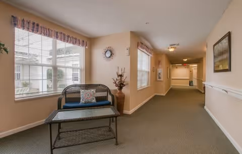 A well-lit hallway in a senior living facility with beige walls and carpeted floor. There is a black wicker loveseat with a blue cushion and a decorative pillow, accompanied by a matching wicker coffee table. A large window with blinds and a valance lets in natural light. The hallway extends into the distance with framed artwork on the walls and a large vase with dried branches near the loveseat.