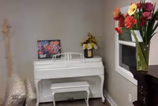 A small white piano with a matching bench is placed against a light gray wall. On top of the piano, there is a colorful floral picture and a vase with yellow flowers. To the left of the piano, there is a wooden coat rack. To the right, a window with white trim is visible, and in front of the window, there is a tall vase with a bouquet of red, yellow, and orange flowers.