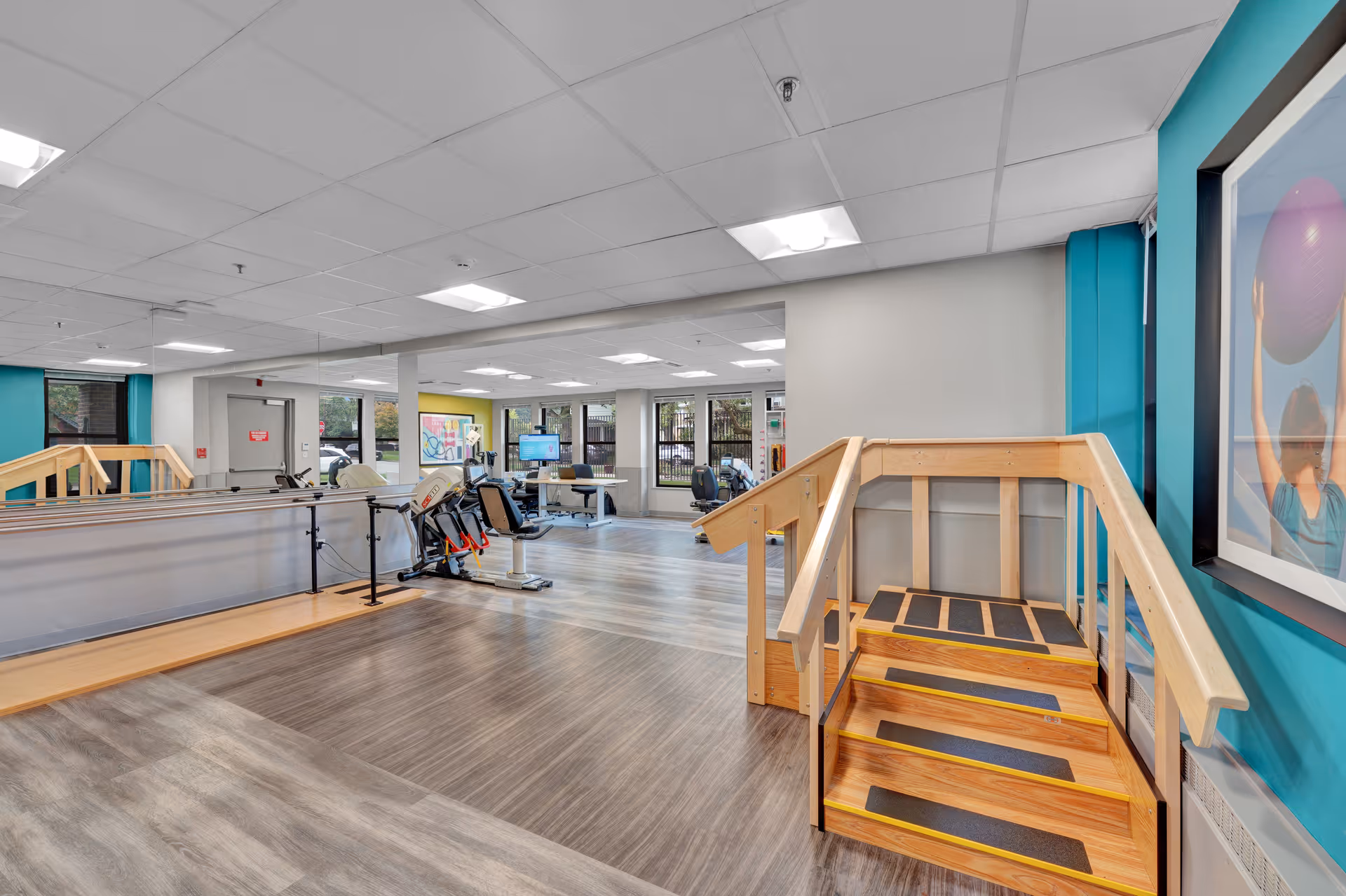 Interior view of a rehabilitation and nursing facility room with wooden handrails and steps for physical therapy, exercise equipment, large windows, and bright overhead lighting.
