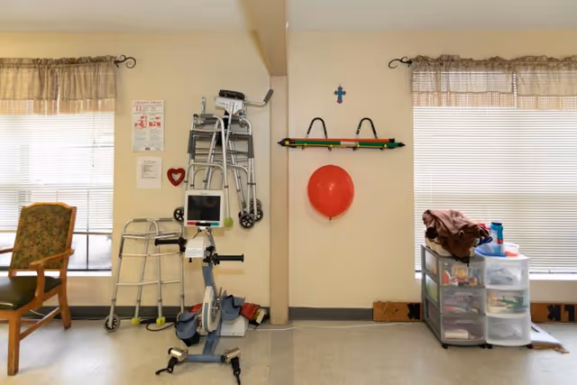 Interior room with medical and mobility equipment including walkers and an exercise bike. There are two windows with blinds and valances, a wooden chair with floral upholstery, a red balloon hanging on the wall, and a plastic drawer unit with various items on top. The walls are light-colored and the floor is a light gray.