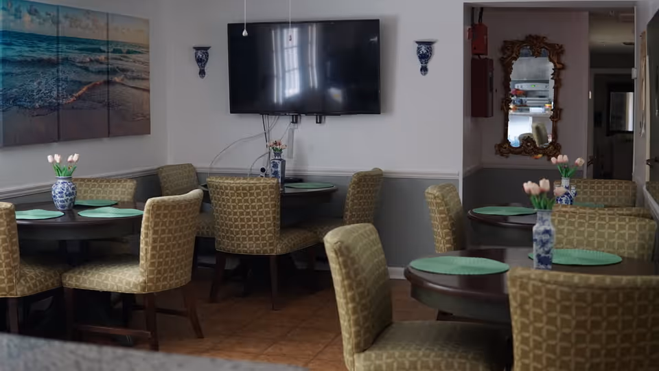 Small dining room with round tables and patterned chairs, each table set with green placemats and blue-and-white vases of tulips, a wall-mounted TV and beach artwork.