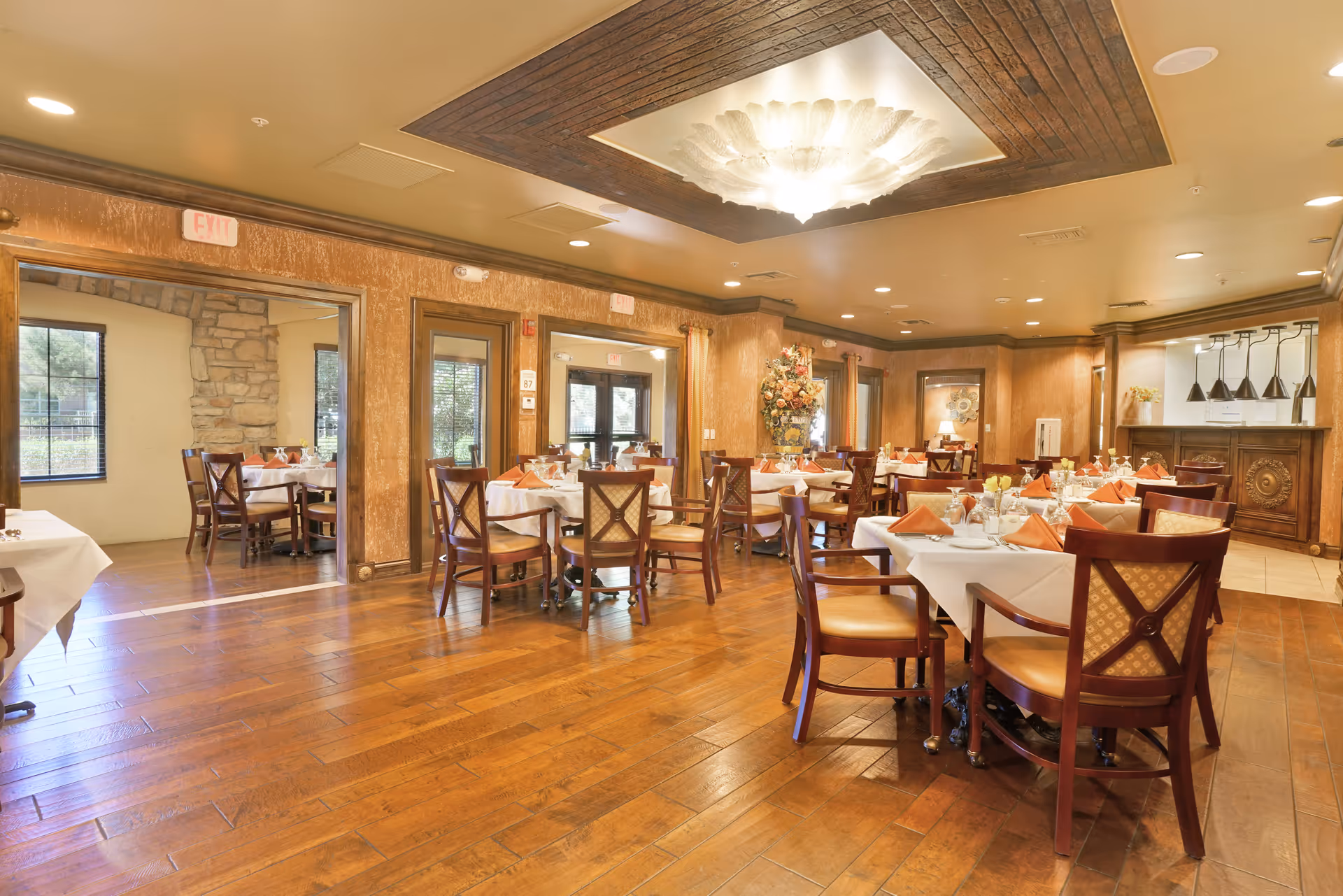 A spacious dining room with wooden floors and multiple tables set with white tablecloths, orange napkins, and glassware. The room features warm-toned walls, a decorative ceiling light fixture, and large windows allowing natural light. There is a floral arrangement in the corner and a serving area with pendant lights in the background.