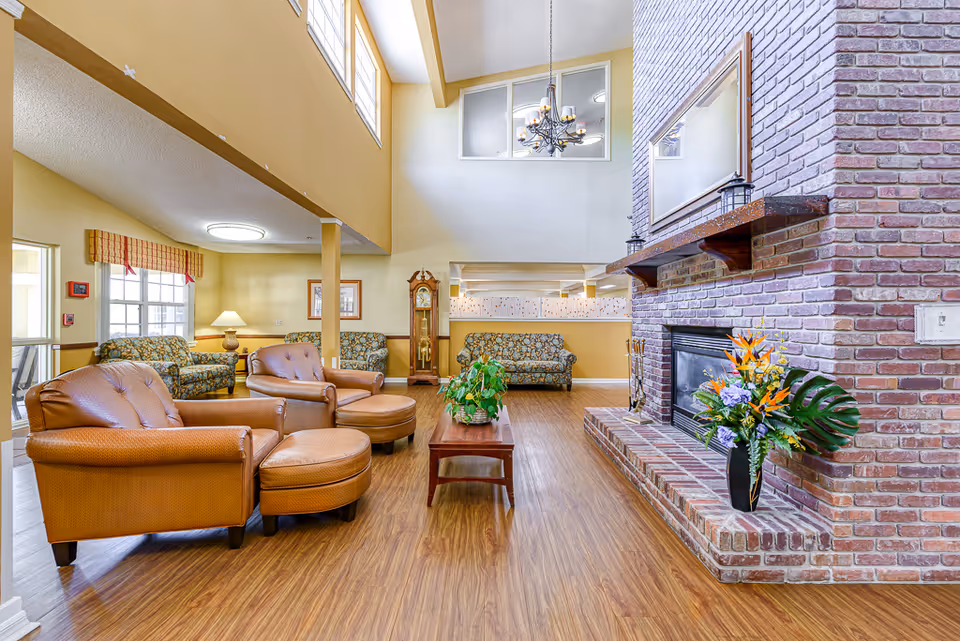 Spacious senior living common room with leather armchairs and ottomans, patterned sofas, a central coffee table with plants, and a large brick fireplace under high ceilings.