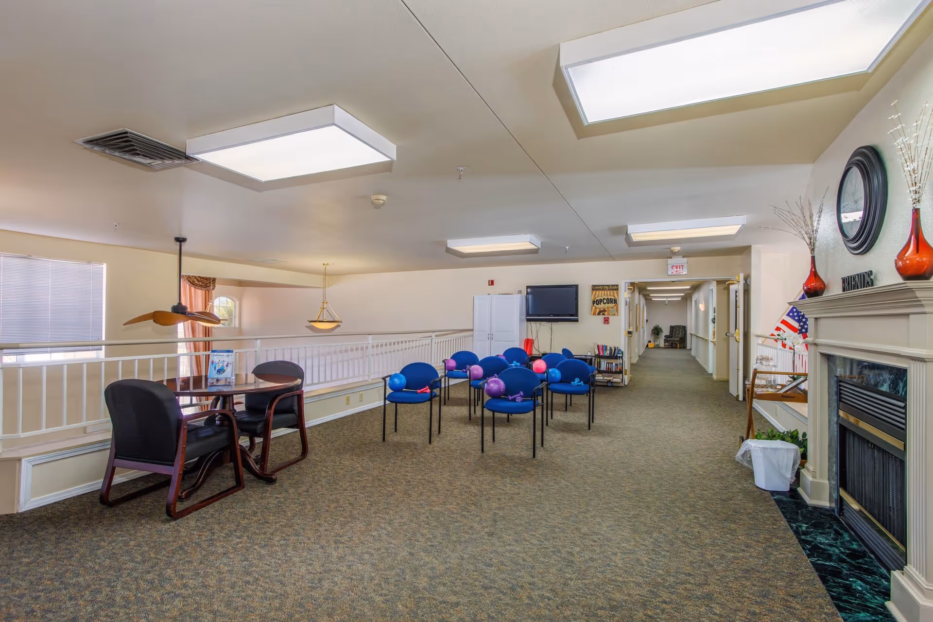 A spacious common area in an assisted living facility with blue chairs arranged in rows, some with purple exercise balls on them. There is a small table with two black chairs on the left side near a railing, and a fireplace with decorative vases and an American flag on the right. The room has beige walls, carpeted floor, and ceiling lights. A TV is mounted on the far wall above a small bookshelf, and a hallway extends from the back of the room.