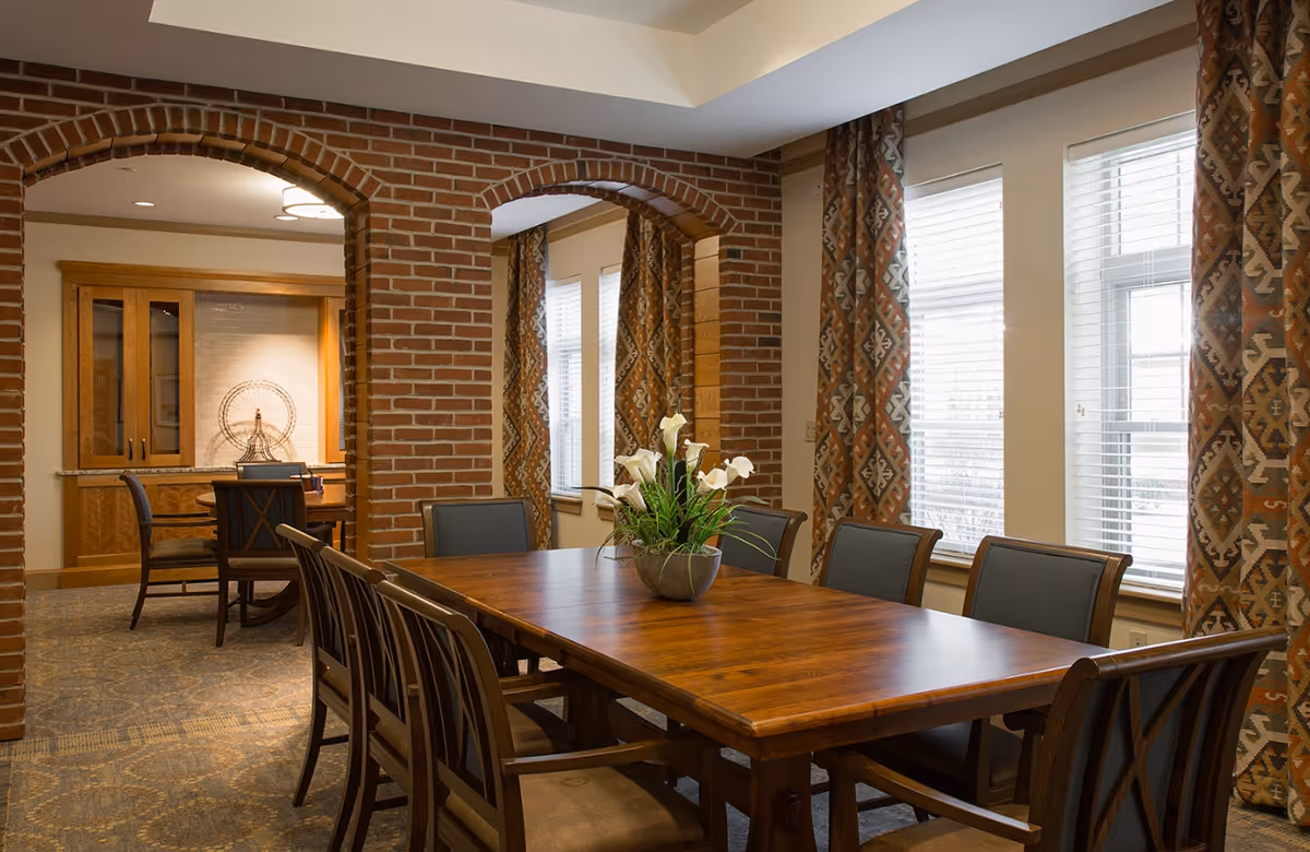 A dining room with a large wooden table surrounded by eight chairs. The room features brick archways, patterned curtains on windows with white blinds, and a decorative centerpiece with white flowers on the table. In the background, there is another smaller table with chairs and a cabinet with glass doors.