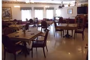 A dining room with multiple square wooden tables and chairs arranged neatly. The room has large windows with valances, framed artwork on the walls, and a television mounted in the corner. The floor is light-colored and shiny, and the room is well-lit with ceiling lights.
