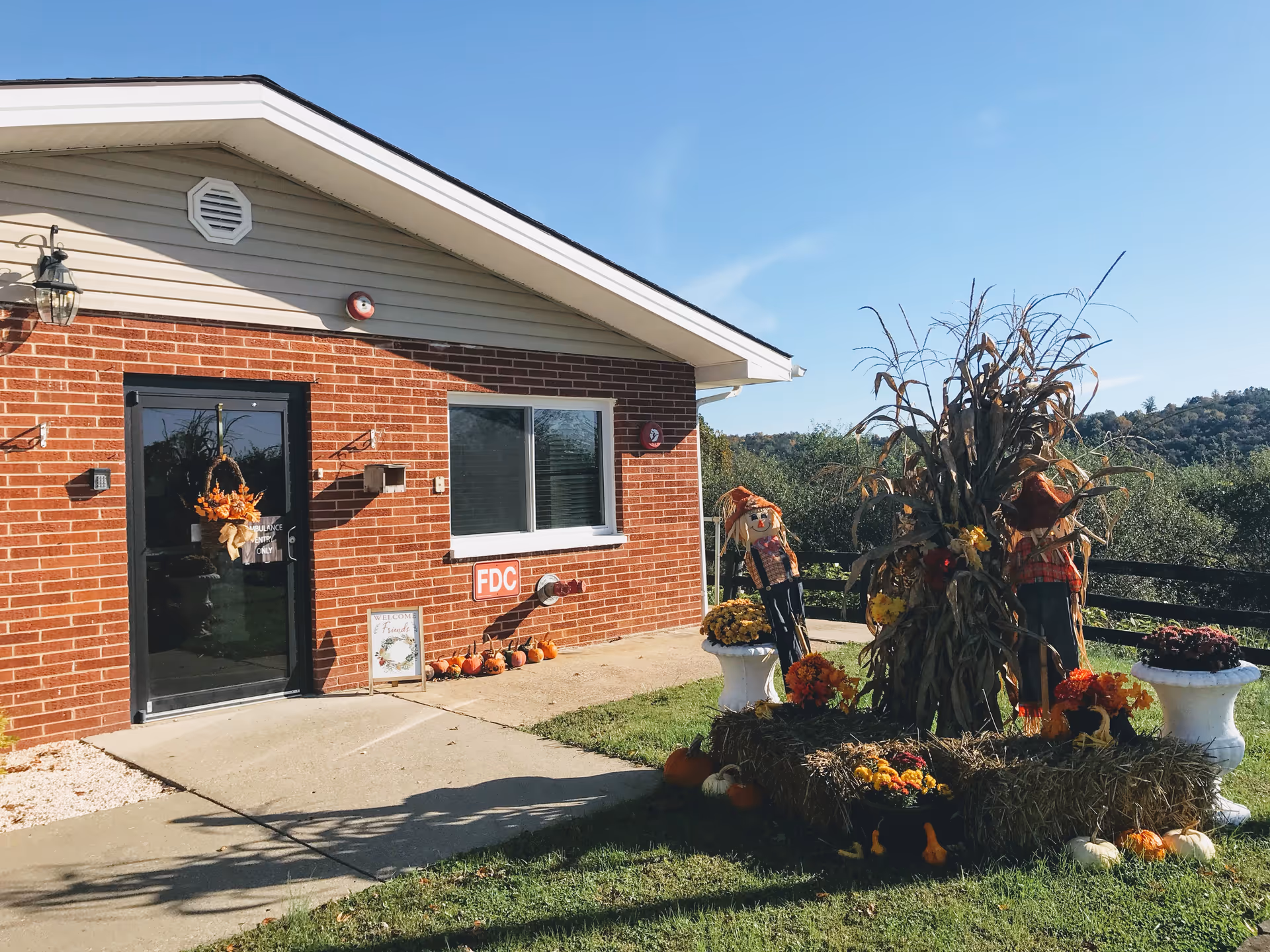 Exterior view of a brick building with a black door decorated with a fall-themed wreath. There is a small sign next to the door and a fire department connection (FDC) on the wall. In front of the building, there is a fall display with hay bales, pumpkins, flowers, corn stalks, and two scarecrows. The scene is set on a sunny day with a clear blue sky and green trees in the background.