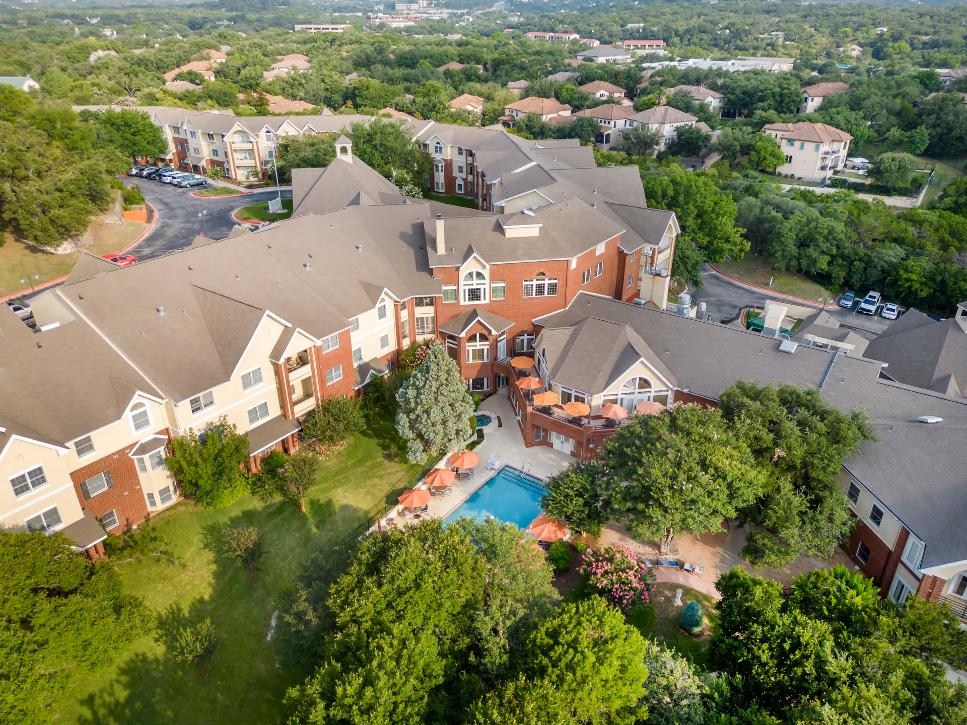 Aerial view of Brookdale Westlake Hills senior living facility showing a large multi-story building with beige and red brick exterior surrounded by trees and greenery. The facility includes an outdoor swimming pool with orange umbrellas and lounge chairs. Parking areas and neighboring residential houses are visible in the background.