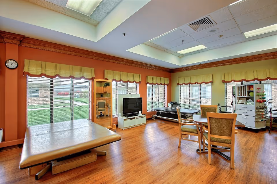 Spacious bright activity room with large windows, exercise tables, a TV, and a table with chairs on wood floors.