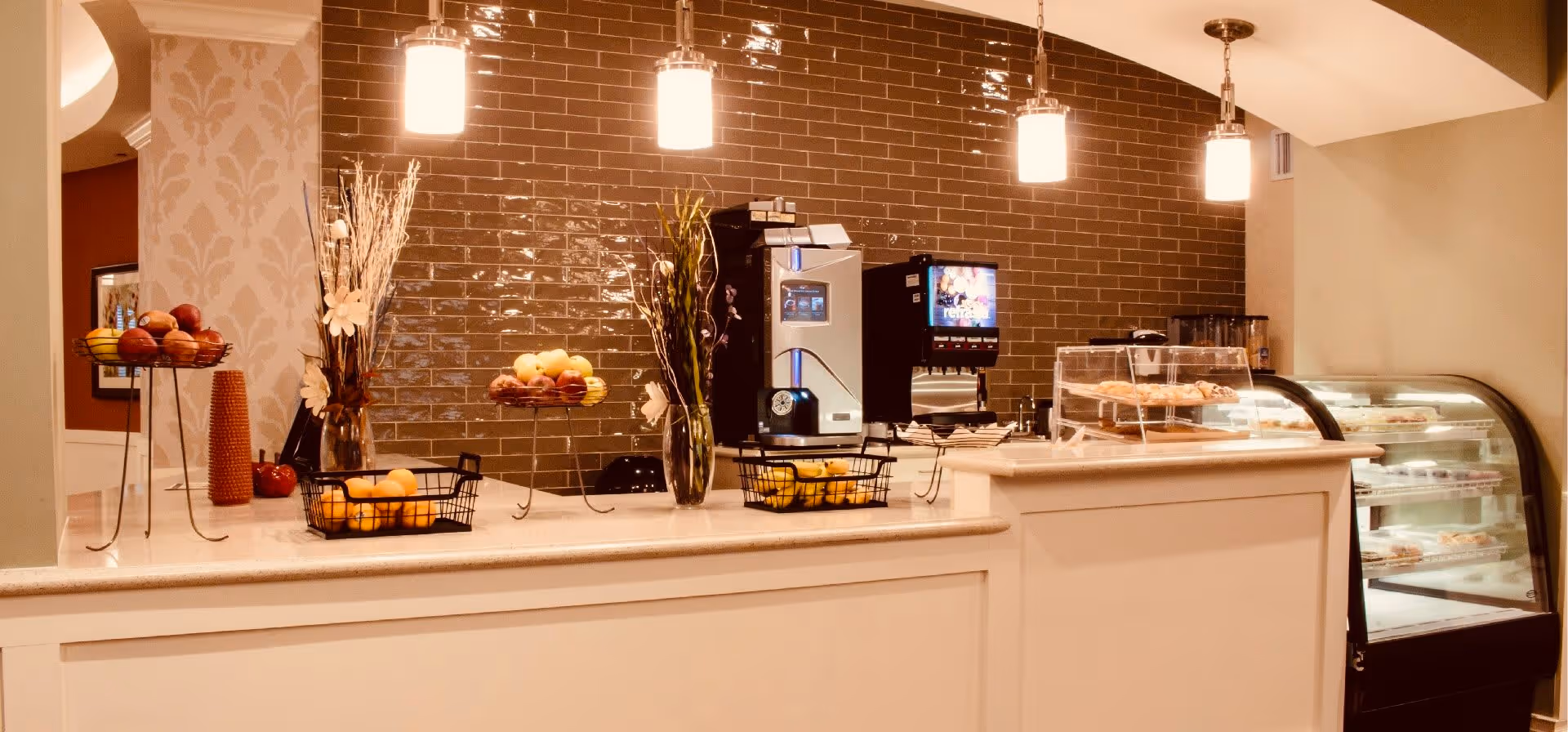 A well-lit counter area with a coffee machine, a soda fountain, baskets of fruit, decorative vases with flowers, and a display case containing pastries. The background features a glossy brown tiled wall and three hanging pendant lights.
