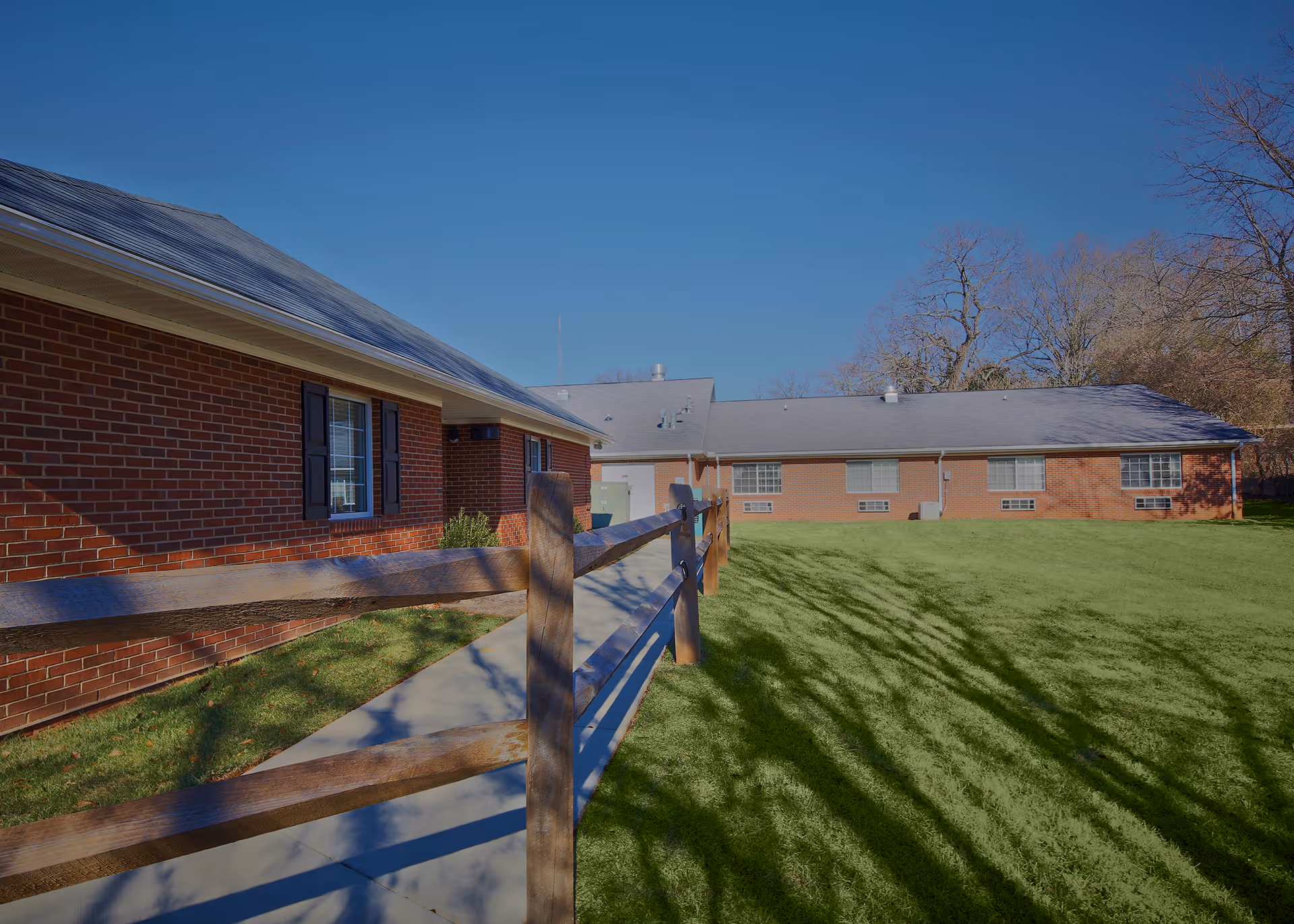 Single-story brick memory care building with a wooden fence, sidewalk, and a green lawn under a clear blue sky.