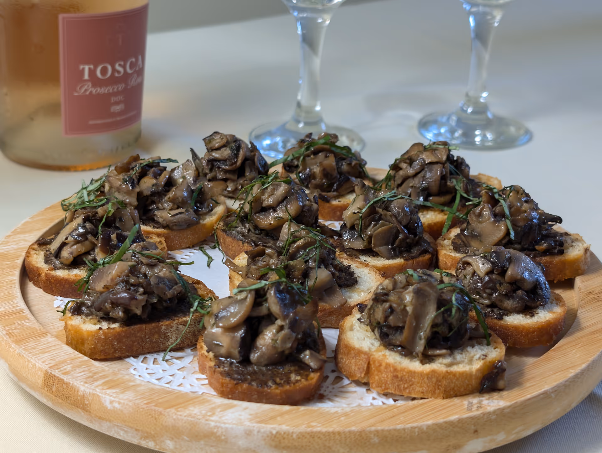 A wooden platter with several pieces of toasted bread topped with sautéed mushrooms and garnished with herbs. In the background, there is a bottle of Tosca Prosecco Rosé and two empty wine glasses.
