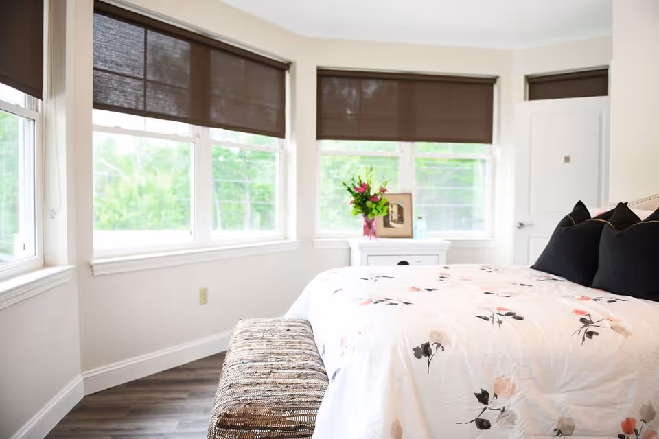 A bright bedroom with large windows covered by brown blinds. The room features a bed with a white floral bedspread and black pillows, a woven bench at the foot of the bed, and a white nightstand with a vase of flowers and a framed picture. The floor is wooden, and the walls are painted white.