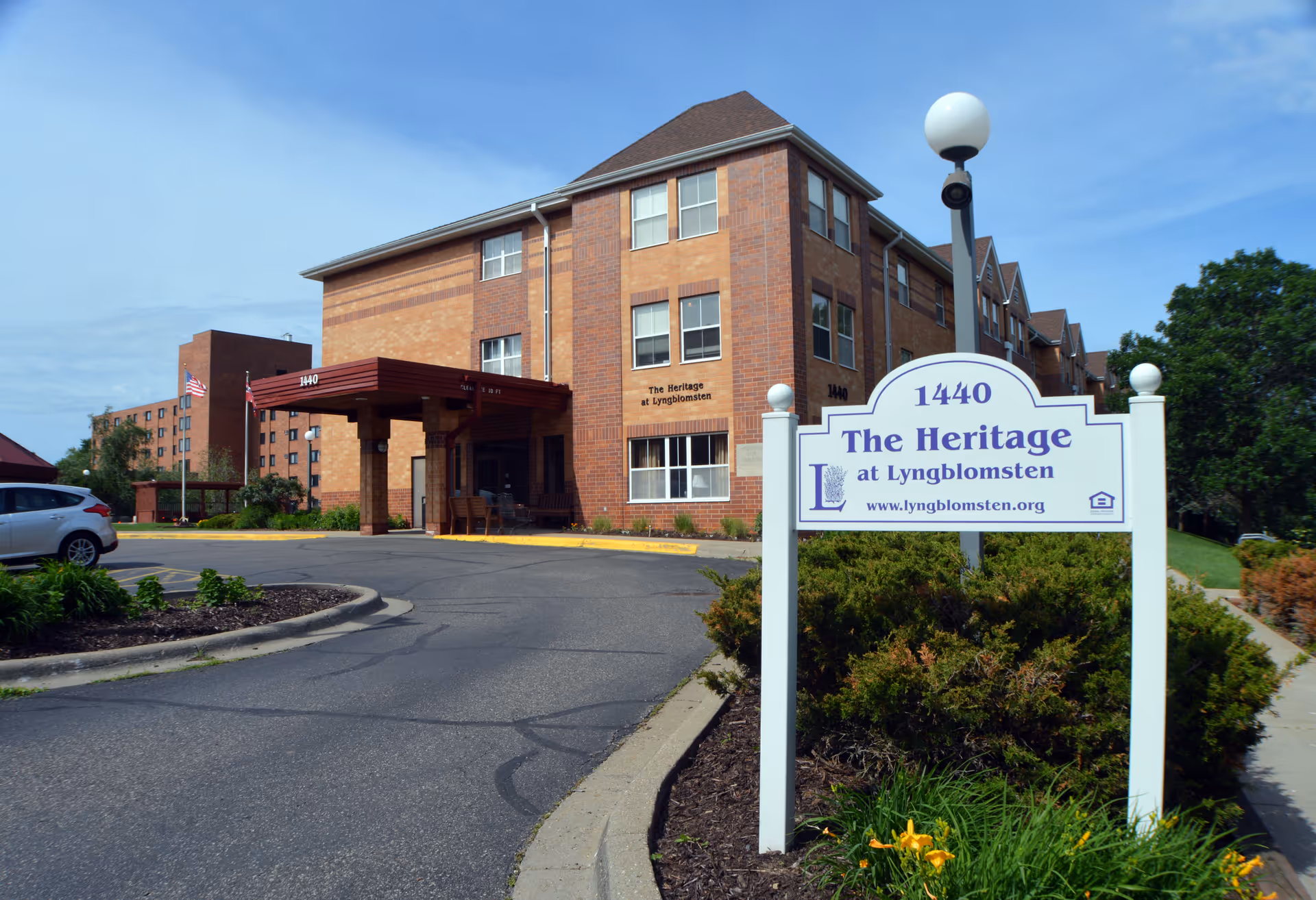 Exterior view of The Heritage at Lyngblomsten senior living facility showing a multi-story brick building with a covered entrance, a driveway, parked cars, and a white sign with the facility's name and website in front of landscaped greenery.