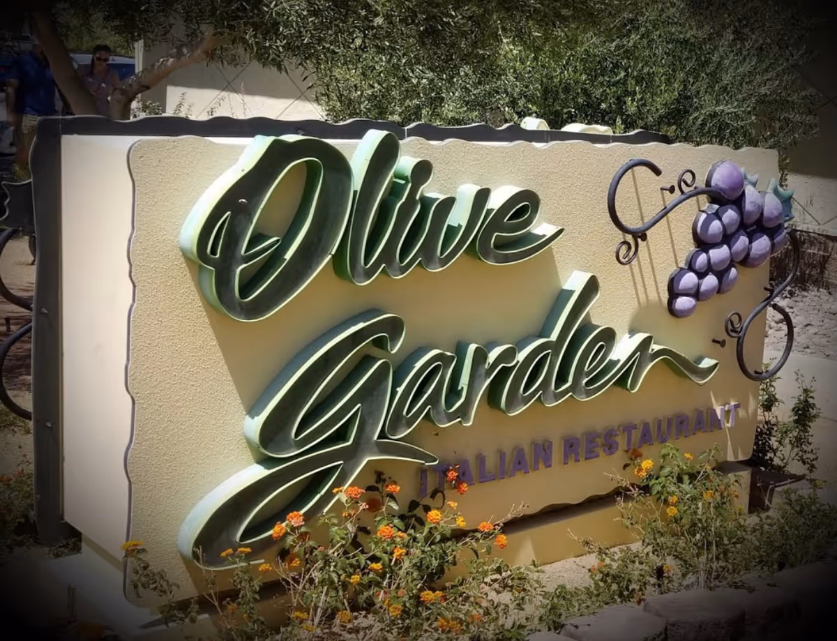 A large outdoor sign for Olive Garden Italian Restaurant with green cursive lettering and a cluster of purple grapes decoration, surrounded by plants and flowers.