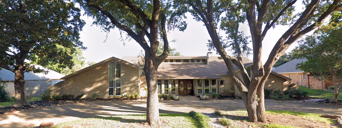 Front exterior view of a single-story building with a sloped roof, large windows, and a central entrance door. The building is surrounded by trees and landscaping with a paved walkway leading to the entrance.