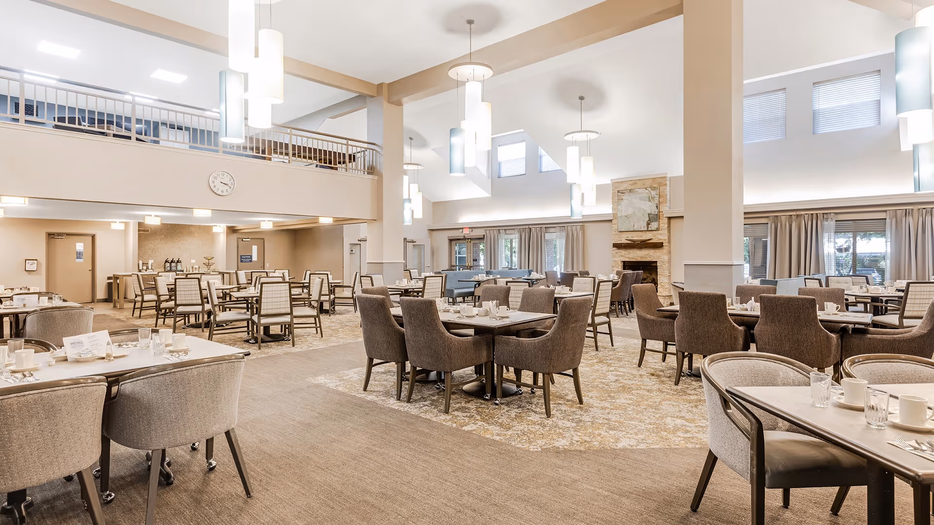 A spacious dining room with multiple tables and chairs arranged neatly. The room features high ceilings with modern hanging light fixtures, large windows with curtains, and a stone fireplace. The decor is neutral with beige and brown tones, and there is a clock on the wall above a doorway in the background.