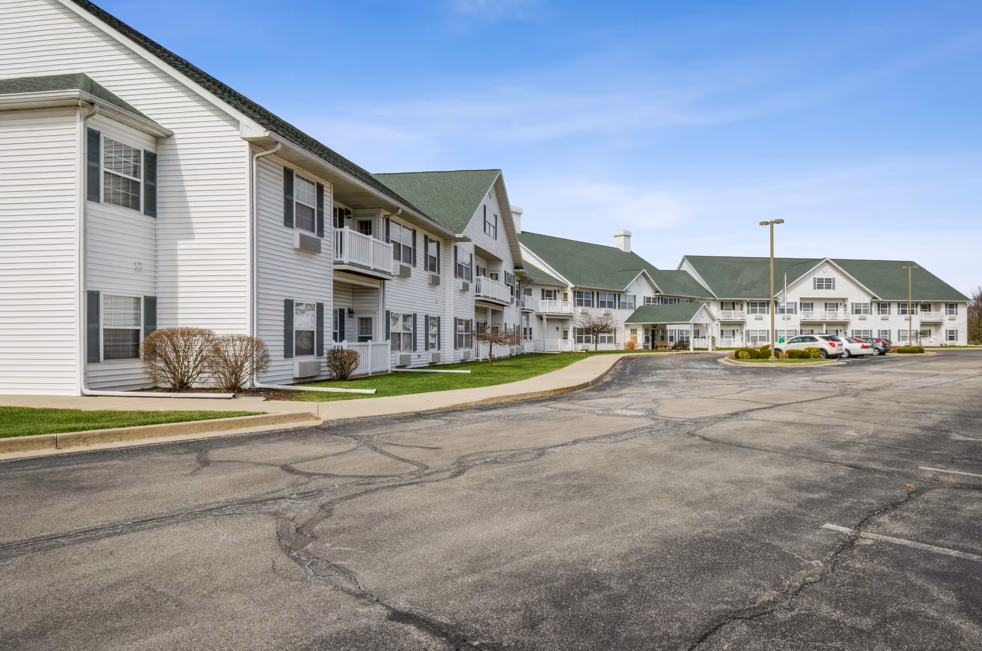 Exterior view of Northwood Manor, a large two-story white building with green roofs, multiple windows, balconies, and a parking lot in front under a clear blue sky.