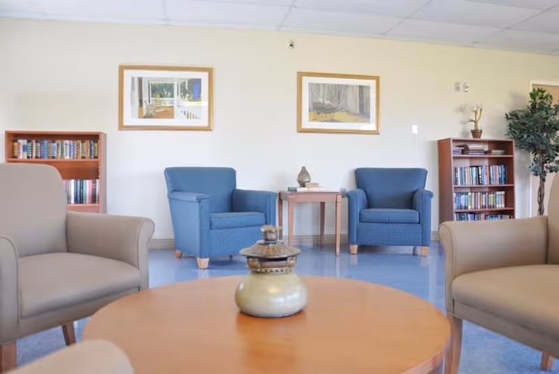 A cozy sitting area in a rehabilitation and nursing center featuring two blue armchairs and two beige armchairs arranged around a round wooden coffee table with a decorative vase. There are two wooden bookshelves filled with books on either side of the room, two framed pictures on the wall, and a potted plant in the corner.