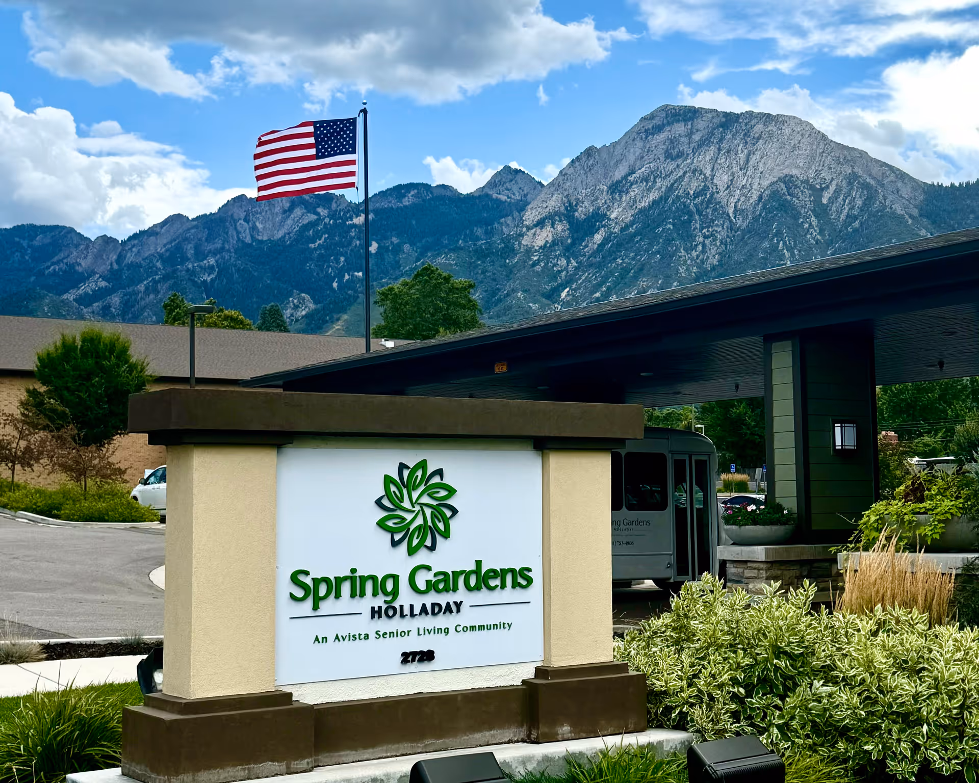 Outdoor view of the entrance sign for Spring Gardens Senior Living Holladay with an American flag on a pole and mountains in the background under a partly cloudy sky.